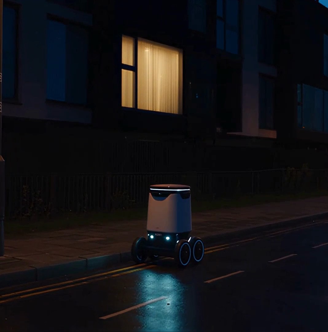 A small delivery robot on a dark, wet street beneath one lit window — the opening of the Folka film.